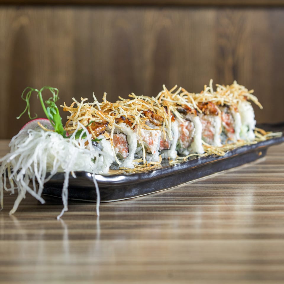 Sushi rolls topped with crispy shredded potatoes, garnishes, and sauce on a black rectangular plate.