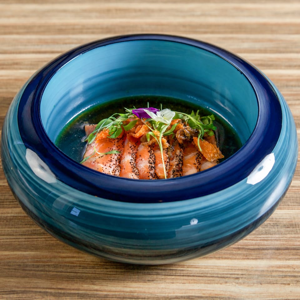 Seared salmon topped with orange relish, microgreens, and edible flowers in a blue ceramic bowl on a wooden table.