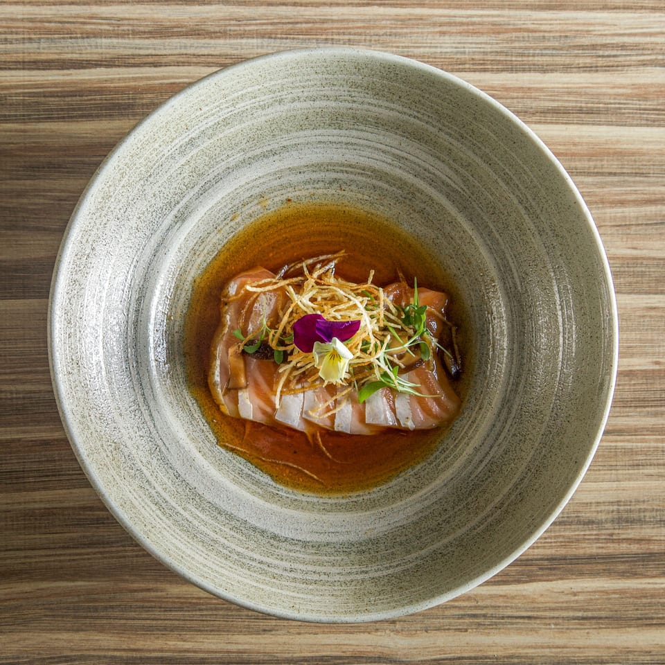Sliced fish in a glossy sauce, topped with crispy shredded garnish, microgreens, and edible flowers in a ceramic bowl.