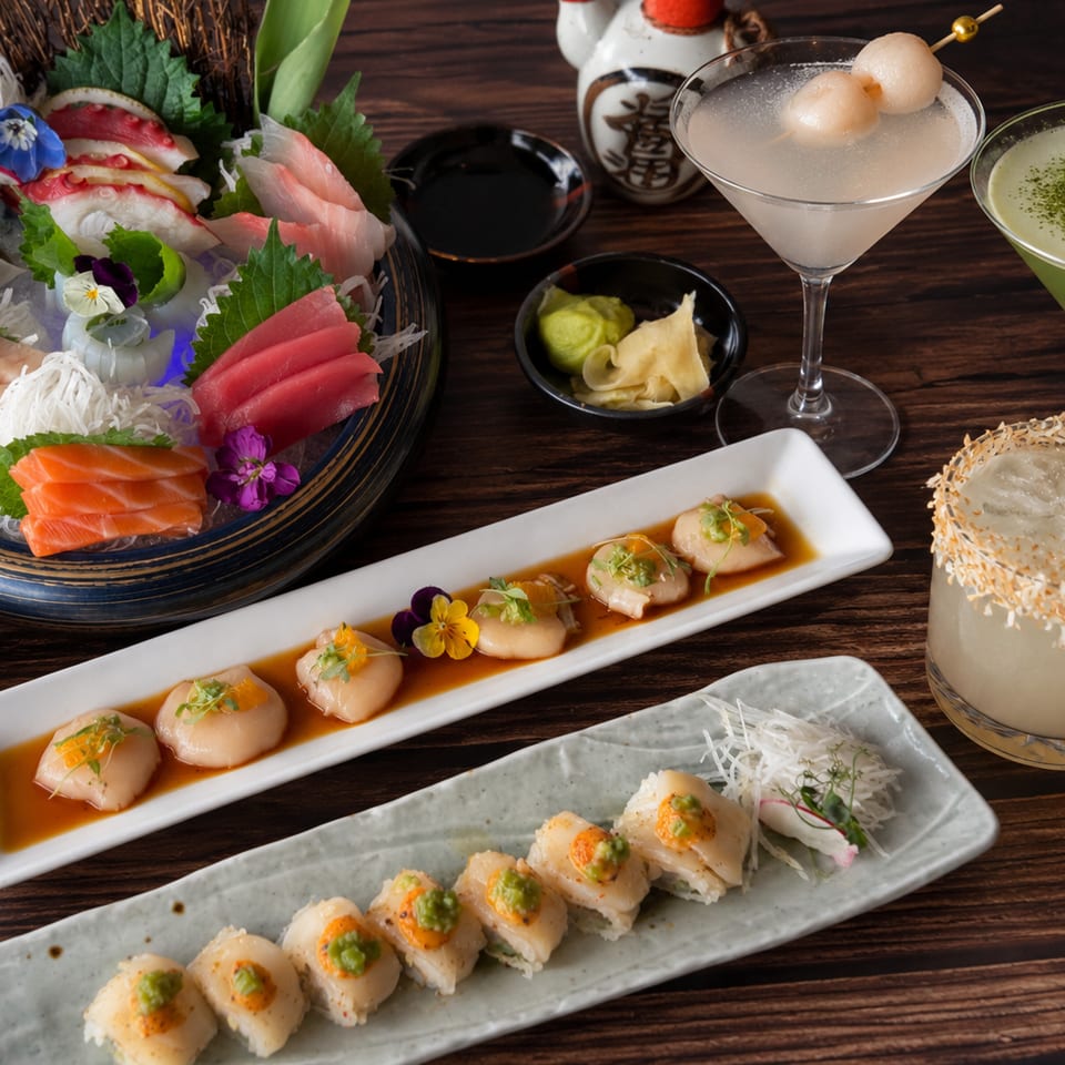 Japanese sushi spread featuring sashimi, scallop dishes, sushi rolls, and three colorful cocktails on a dark wooden table.