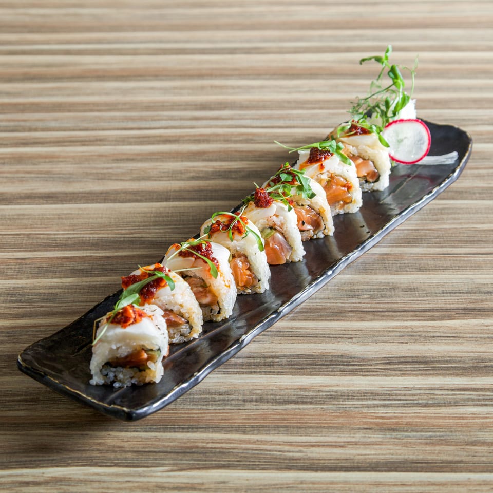 Sushi rolls topped with spicy sauce and fresh herbs on a long black plate, garnished with radish, on a wood-grain table.