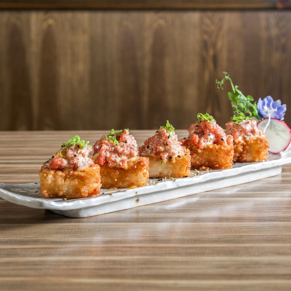 Crispy sushi bites topped with spicy sauce, green onions, and sesame seeds on a white platter with radish garnish.