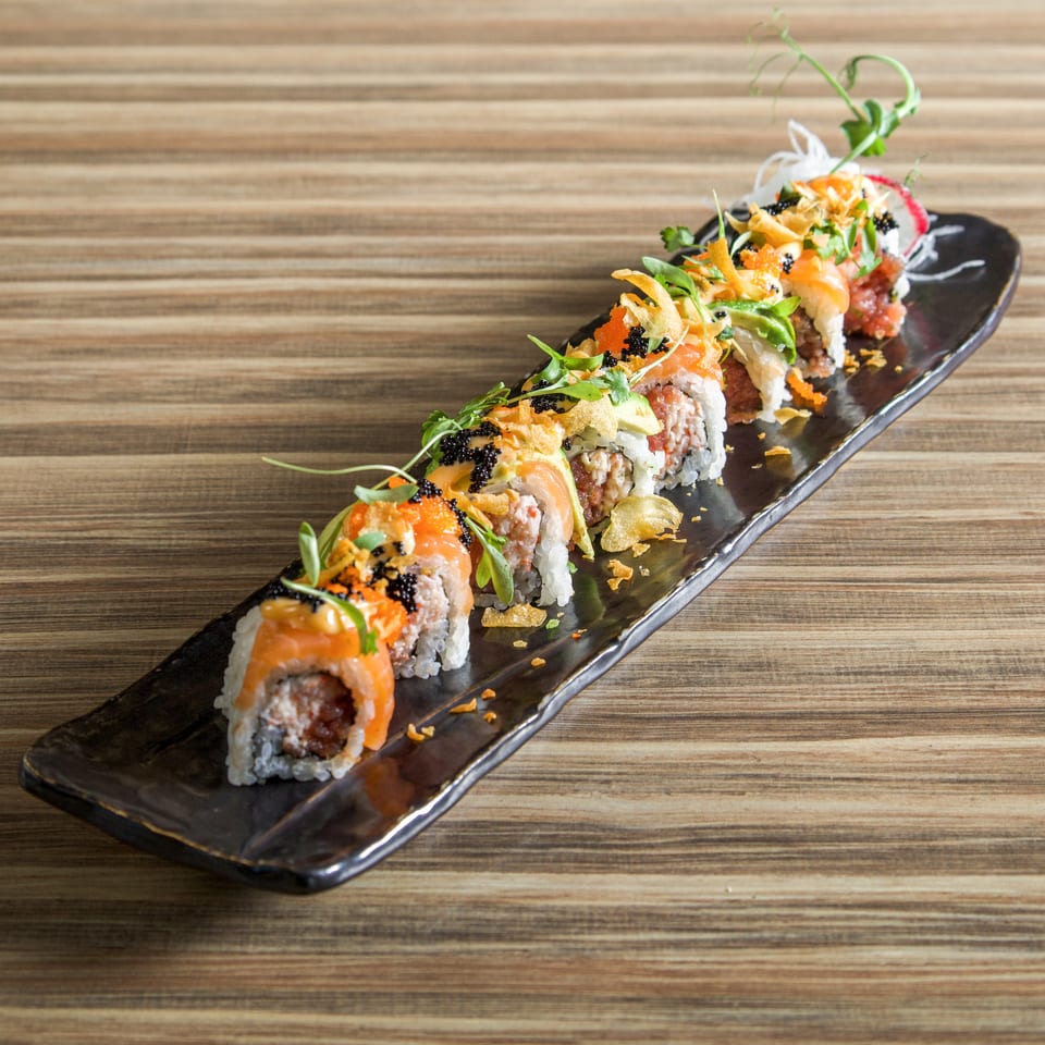 Colorful sushi rolls topped with salmon, avocado, roe, crispy flakes, and herbs on a black platter over a wooden table.