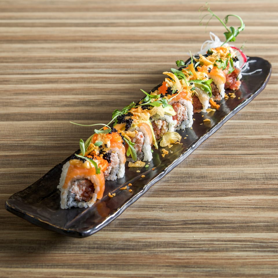 Colorful sushi rolls topped with salmon, avocado, roe, crispy garnish, and herbs arranged on a black platter.