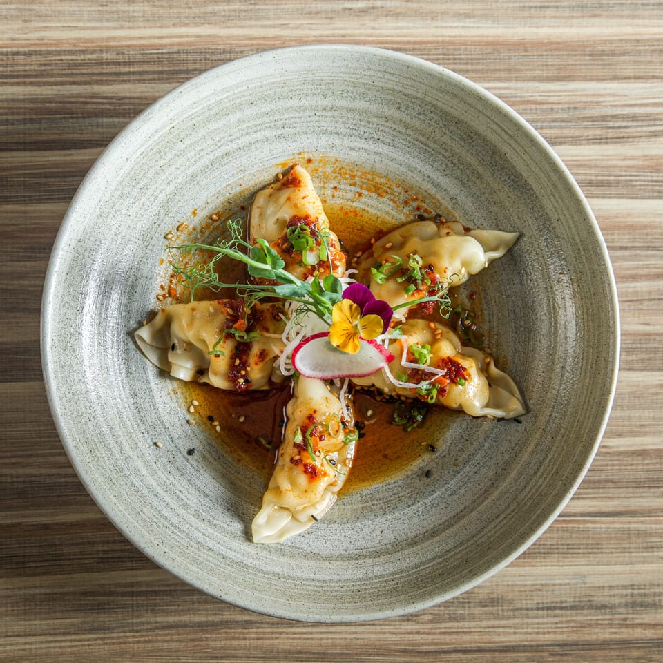 Steamed dumplings coated in chili oil, sesame seeds, and scallions, garnished with microgreens and edible flowers on a ceramic plate.