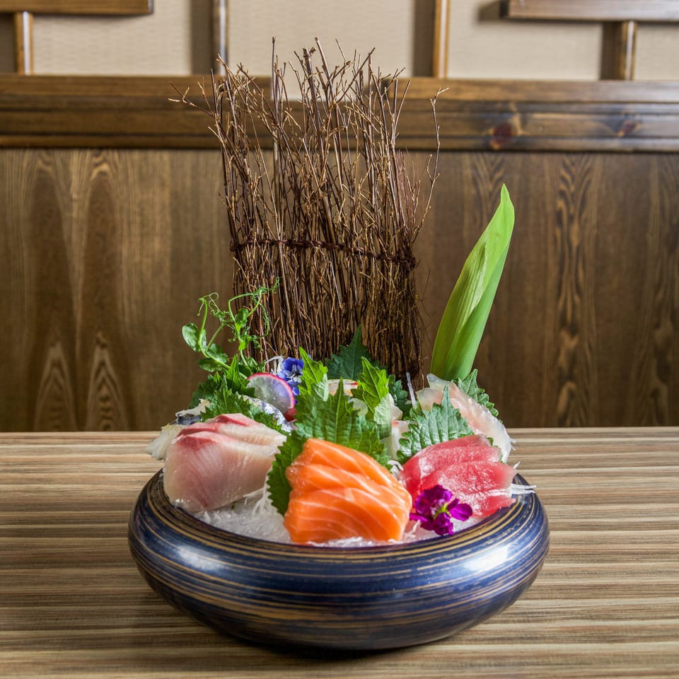 Japanese sashimi platter with salmon, tuna, white fish, herbs, edible flowers, and decorative greenery on a wooden table.