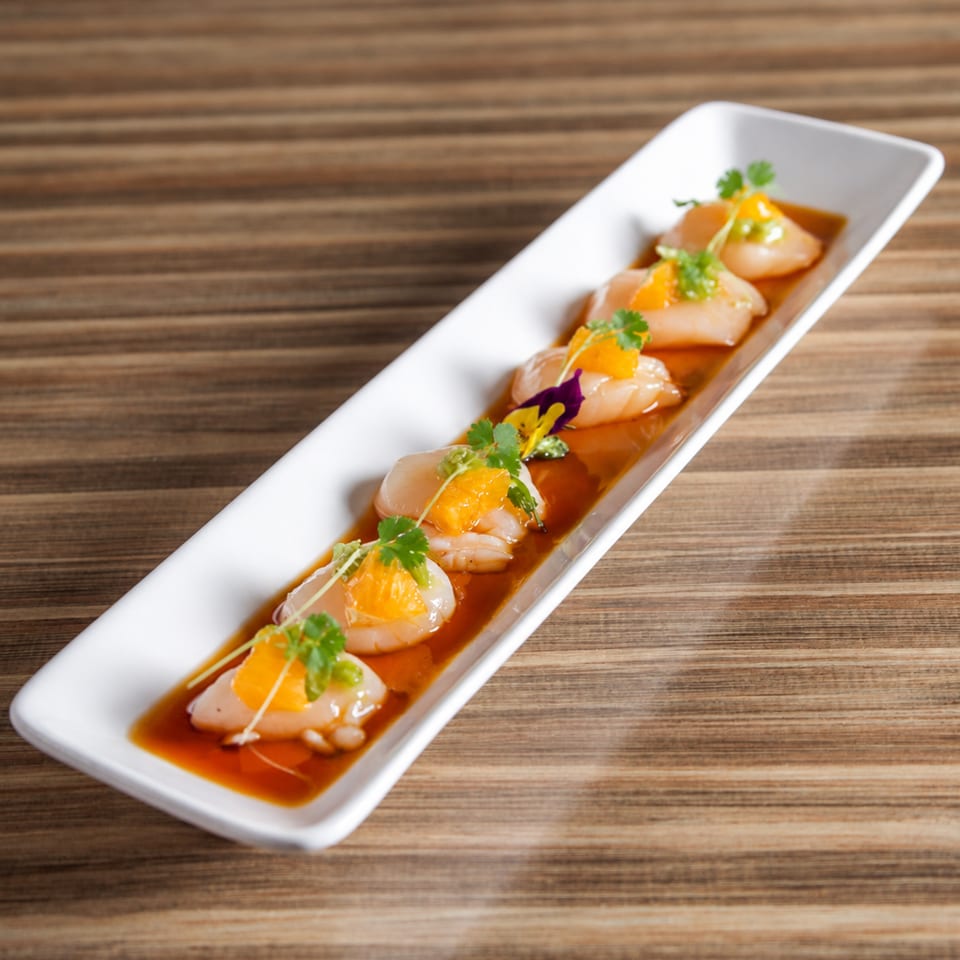 Five scallops topped with orange garnish and herbs, arranged in sauce on a long white platter over a wooden table.