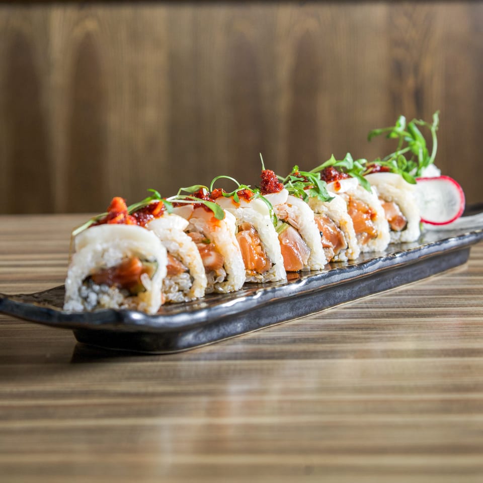 Colorful sushi rolls topped with spicy sauce and greens on a black rectangular platter atop a wooden table.