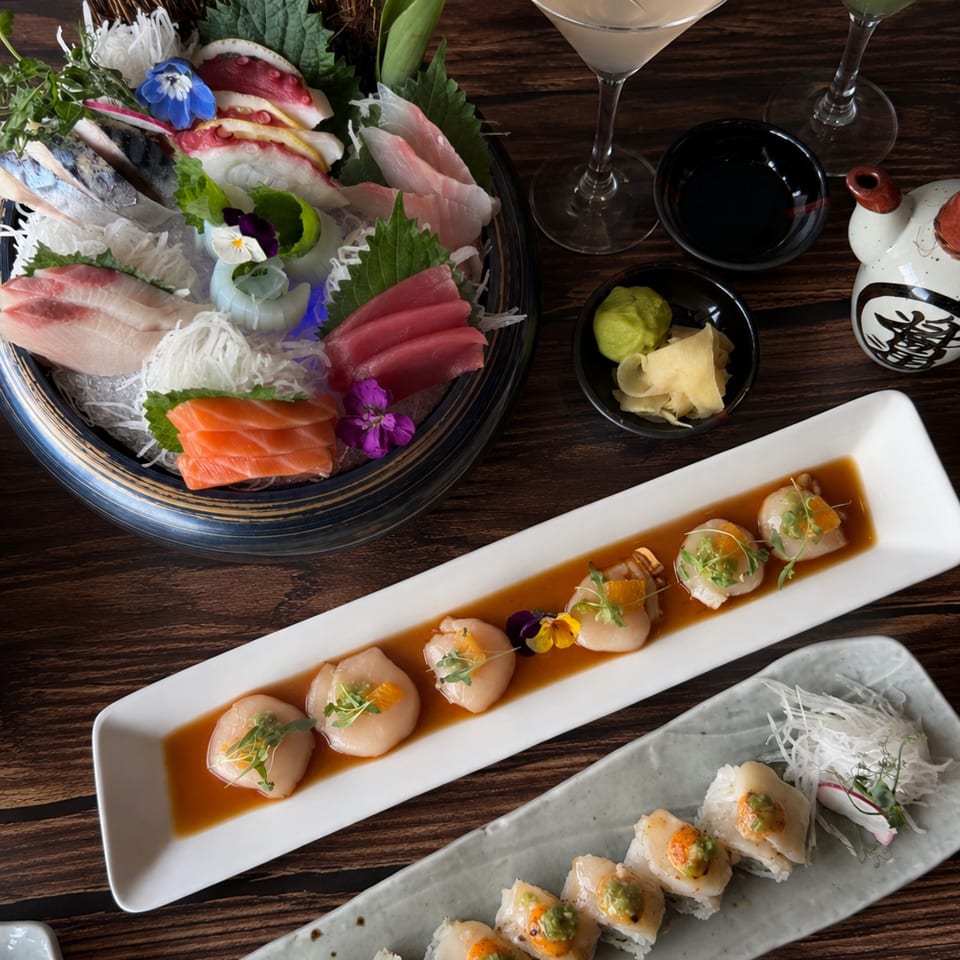 Japanese sushi spread with sashimi, scallop dishes, rolls, dipping sauces, cocktails, and decorative garnishes on a dark wooden table.