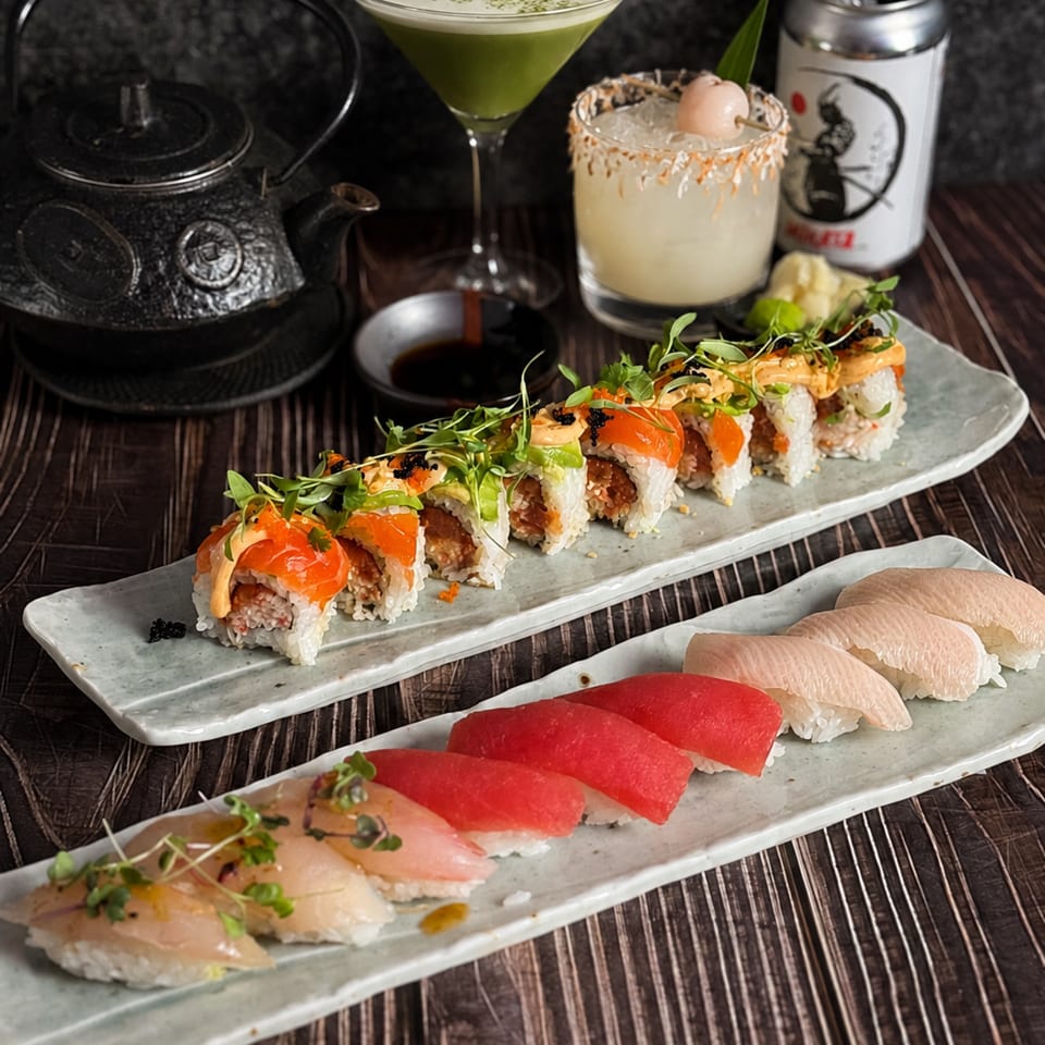 Assorted sushi rolls and nigiri served on ceramic platters with cocktails, tea, and a canned drink on a wooden table.