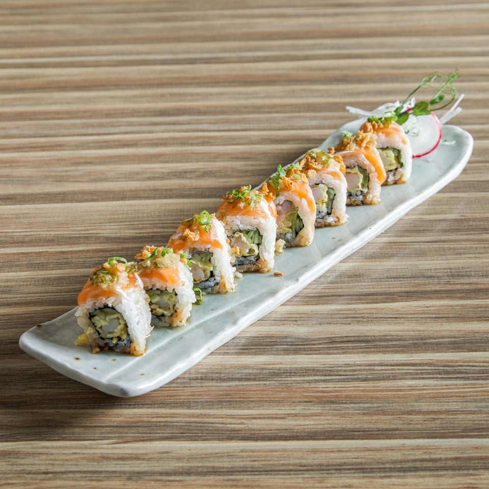 Sushi rolls topped with salmon, crispy garnishes, and scallions on a long white plate atop a wooden table.