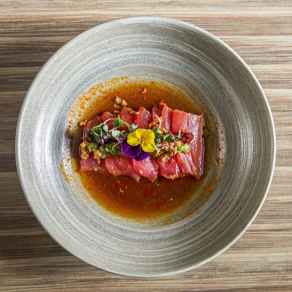 Sliced seared tuna topped with microgreens and edible flowers, served in a spicy sauce on a ceramic plate.