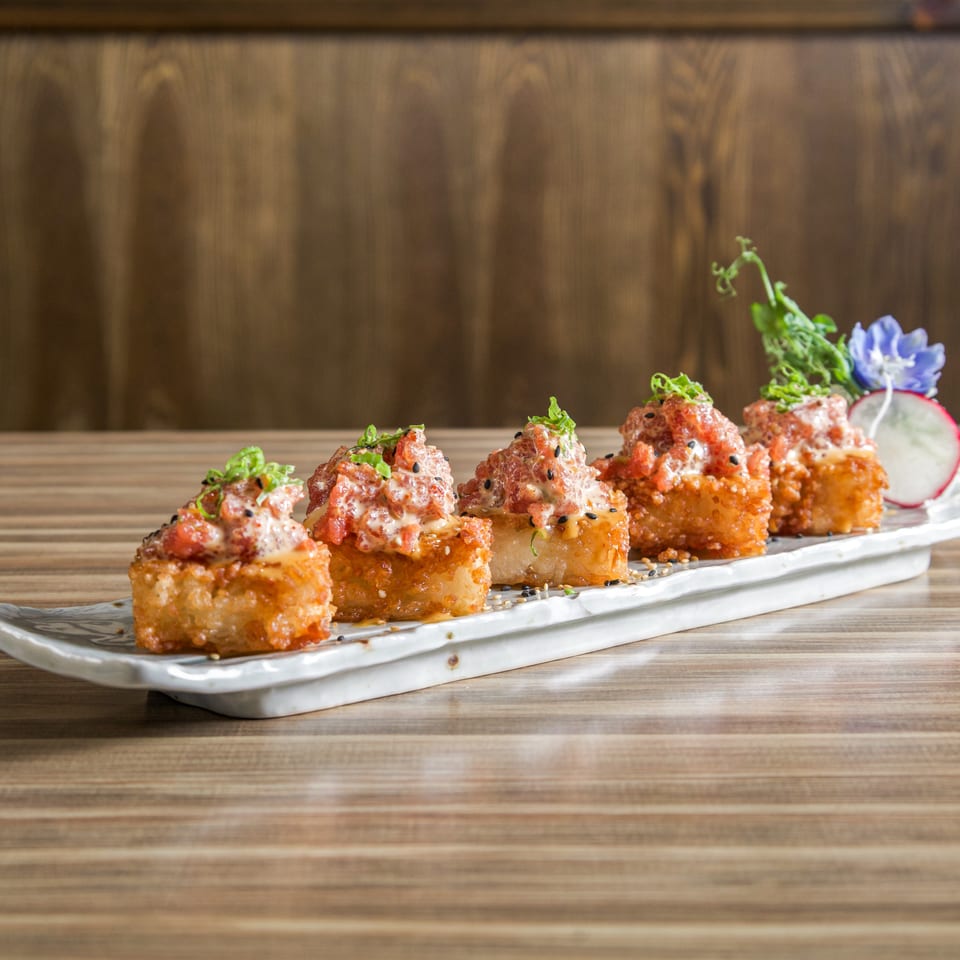 Crispy sushi bites topped with spicy sauce, scallions, sesame seeds, and garnishes on a white rectangular platter.