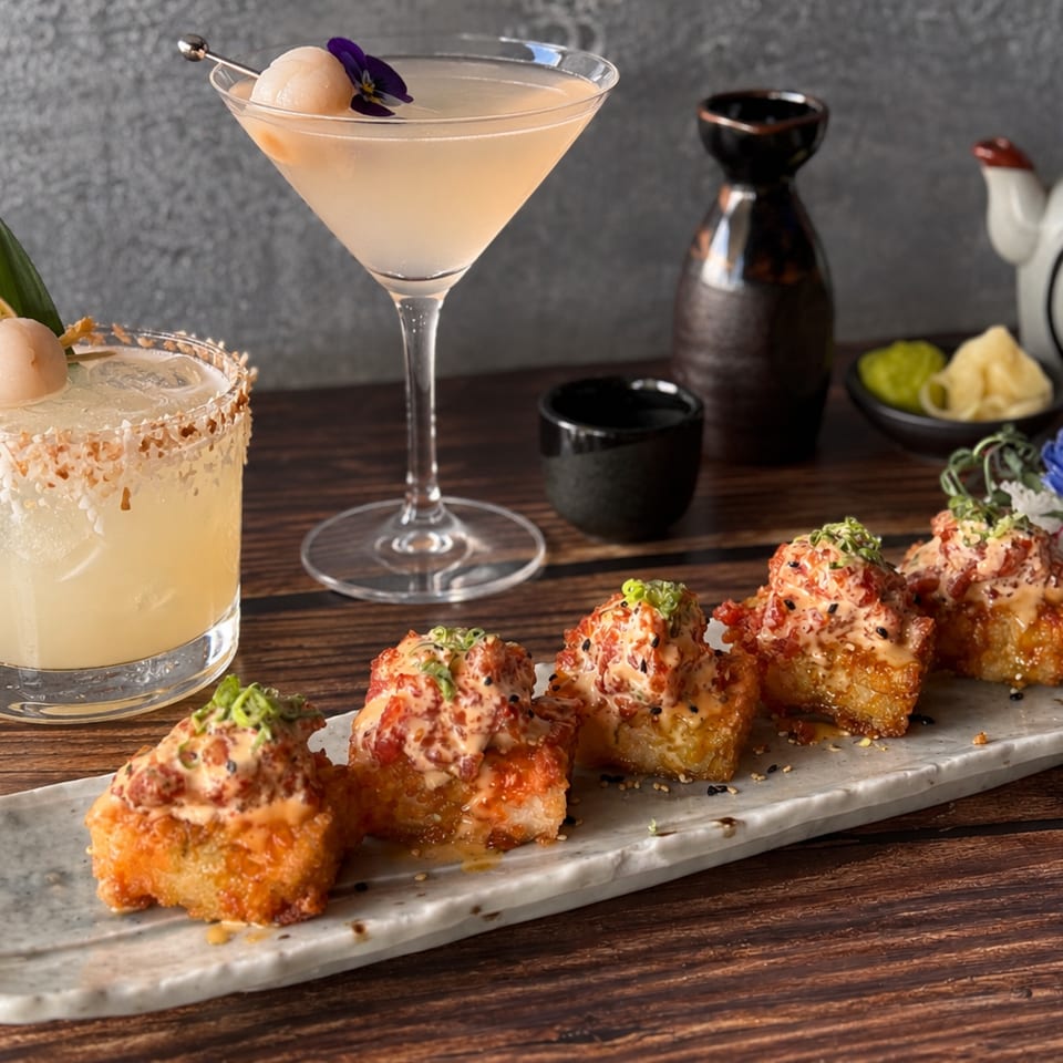 Crispy sushi bites with spicy sauce accompany two lychee-garnished cocktails, sake vessels, and dipping condiments on a wooden table.