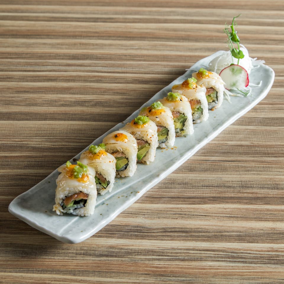 Sushi rolls topped with seared fish and green garnish on a long white plate with radish and herbs.
