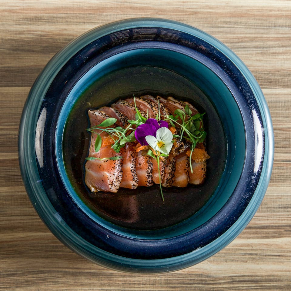 Sliced seared fish topped with microgreens and edible flowers in a dark blue bowl on a wooden table.