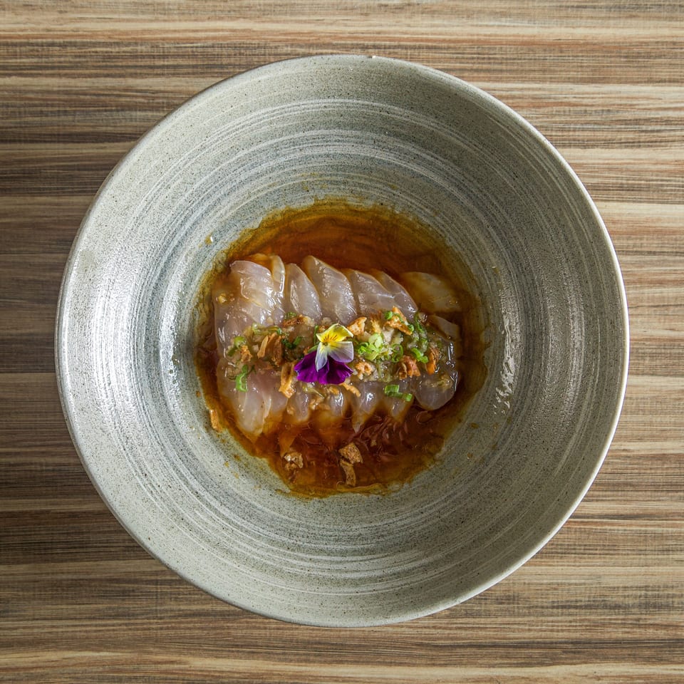 Thinly sliced fish arranged in a bowl of amber sauce, topped with scallions, crispy garnish, and an edible flower.